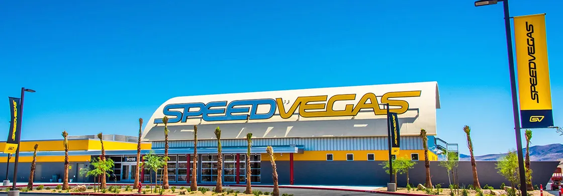 Exterior of Speed Vegas building on a sunny day, featuring a large yellow and blue sign. Tall palm trees line the sidewalk, creating an inviting atmosphere.